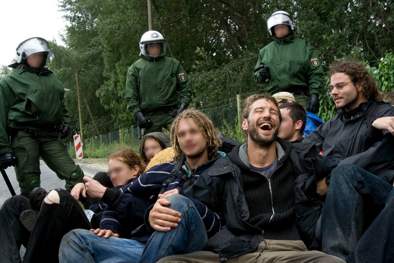 Cops umzingeln eine am Boden sitzende, untergehakte Gruppe von Aktivisten. In der Mitte davon sitze ich und muss aus irgendwelchen Gründen lachen. Neben mir mein alter Freund Alexis Passadakis.