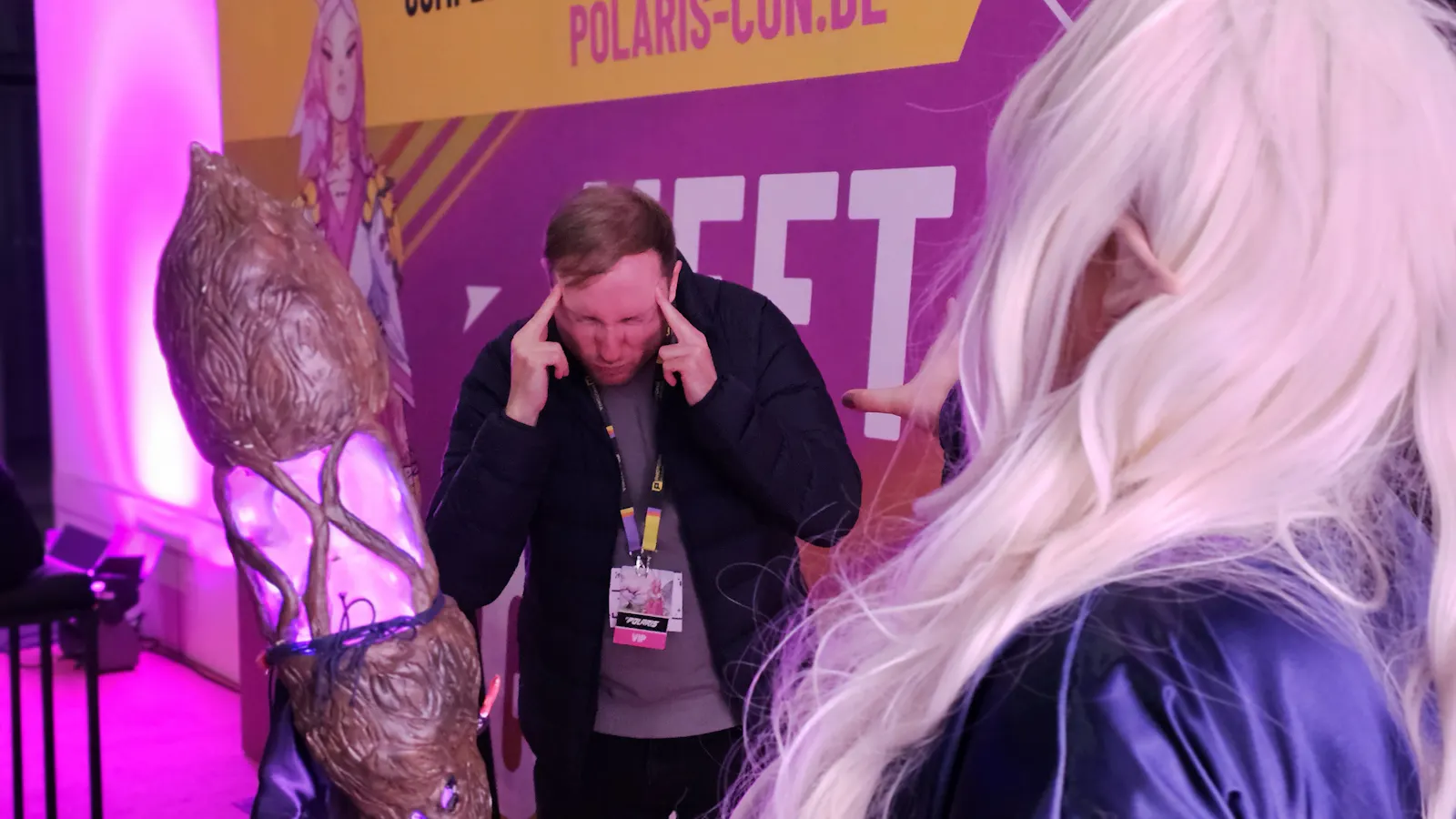 Kelly hält sich beide Zeigefinger an die Schläfen. Im Vordergrund ist von hinten ein Cosplay von Lady Uriella zu erkennen.