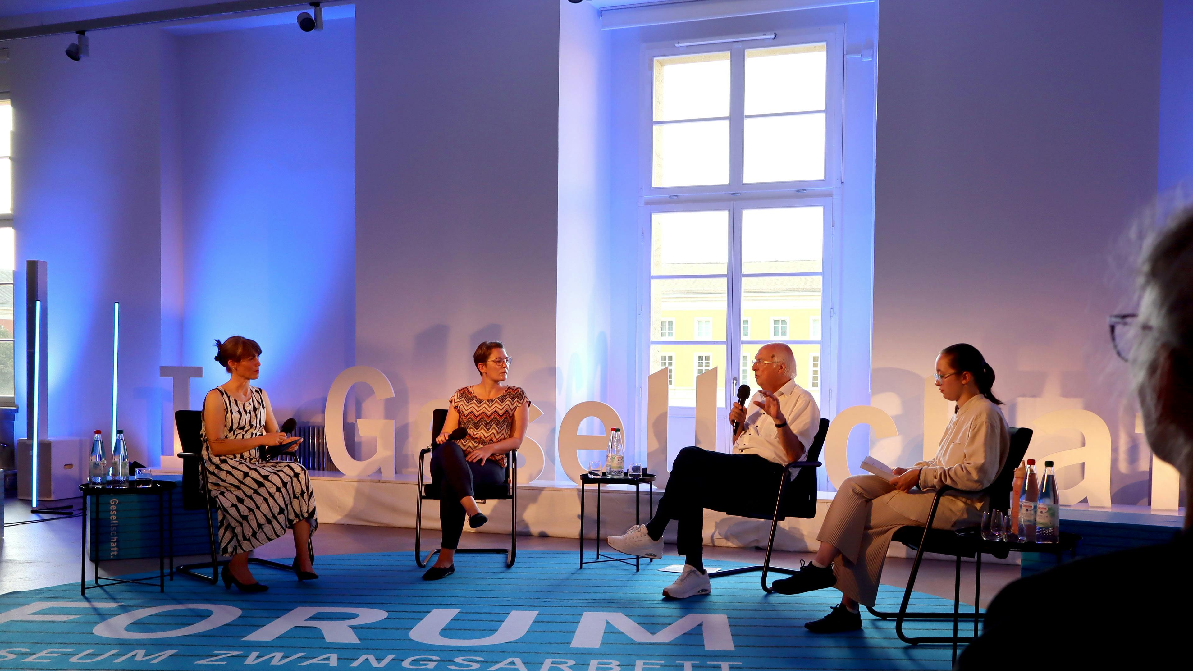 In einem Saal mit hohen Fenstern sitzen vier Menschen auf Stühlen im Halbkreis, sie halten Mikrofone in ihren Händen. Im Hintergrund ist der Schriftzug "In Gesellschaft" zu erkennen. Auf dem Boden liegt ein blauer Teppich mit schwarzen Queerstreifen. Darauf steht in weißer Schrift "Forum Museum Zwangsarbeit Weimar". Am rechten Bildrand ist das Gesicht einer Person aus dem Publikum zu erkennen.