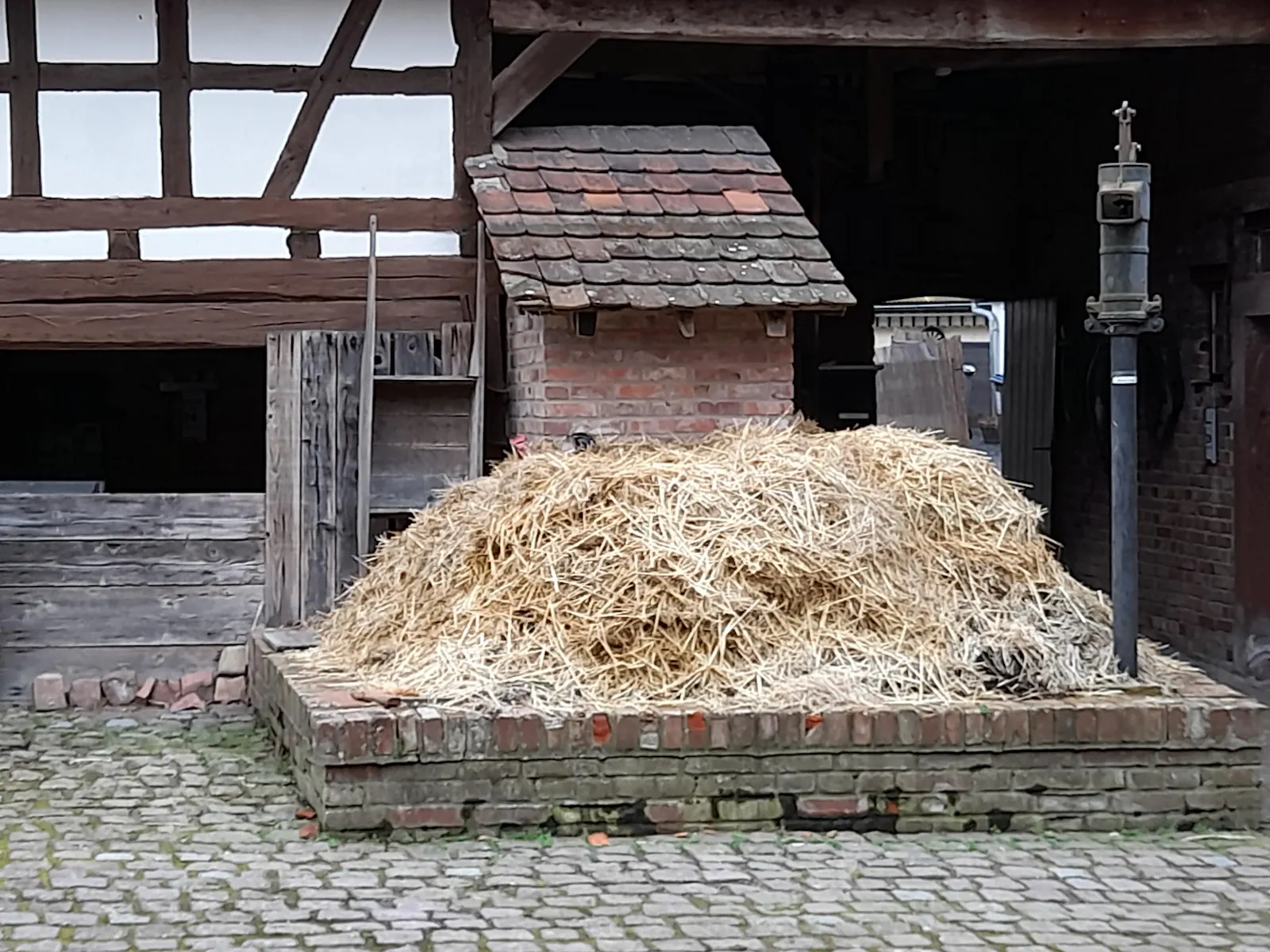 In the courtyard of our farm museum: a dung pile in front of the outhouse. The cast-iron slurry pump rises up on the right. A wooden wheelbarrow for the manure leans on the left at a half-timbered wall.