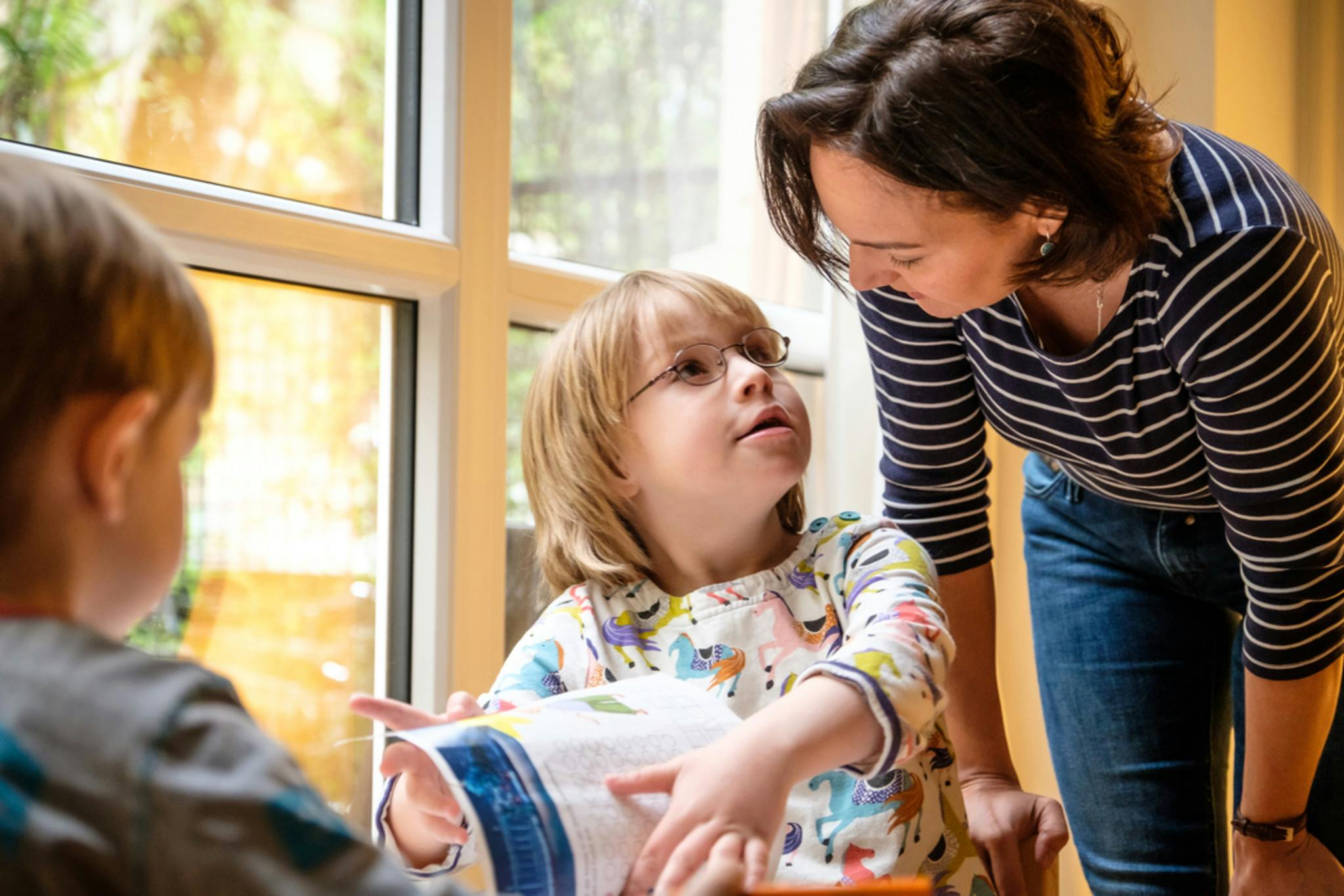 Zwei Kinder sitzen an einem Tisch am Fenster, eine Person beugt sie zu einem der Kinder herunter, das der Person etwas erzählt.