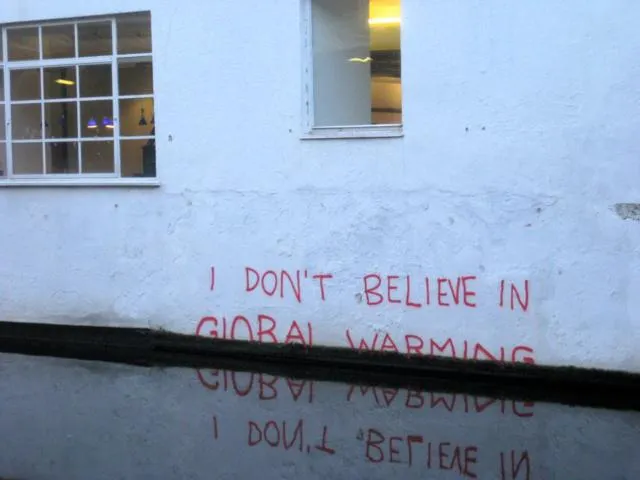 An einer Hauswand steht: I don`t believe in global warming. = Ich glaube nicht an globale Erwärmung. Die Schrift spiegelt sich im Wasserspiegel des Hochwassers, das von globaler Erwärmung verursacht wird.