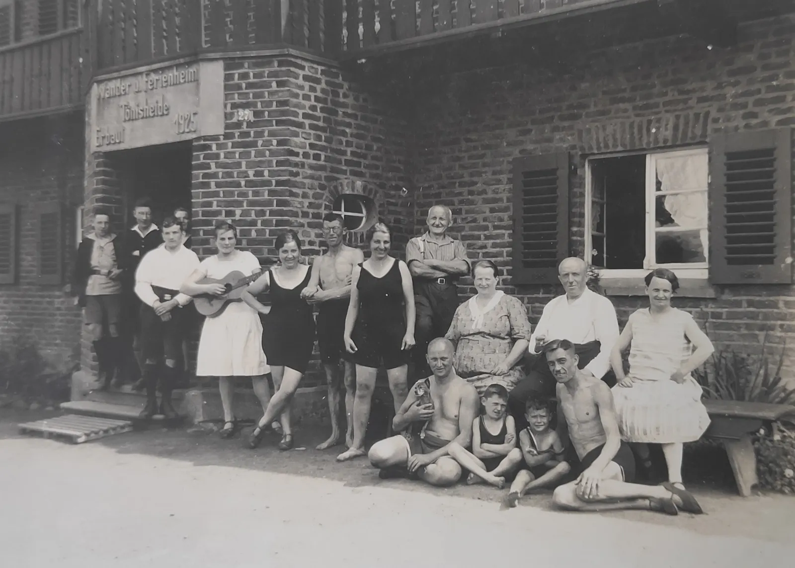 Schwarz-Weiß Foto einer Familie aus 16 Personen in legerer Kleidung vor einem Ziegelbau im Eingangsbereich draußen im Sommer.
