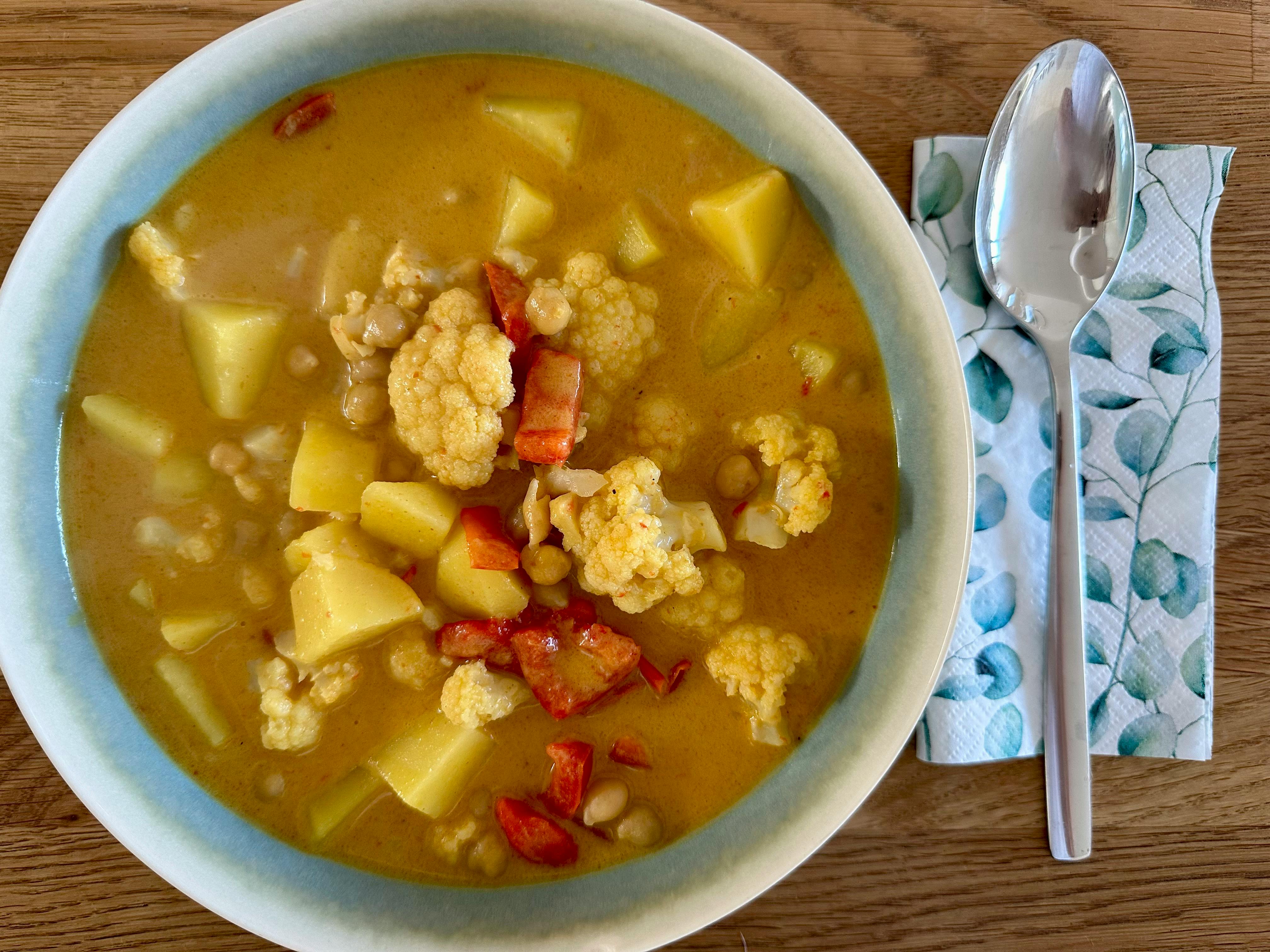 Steingutteller mit einem Curry aus Kartoffeln, Blumenkohl und Paprika. Daneben eine Serviette und ein großer Löffel.