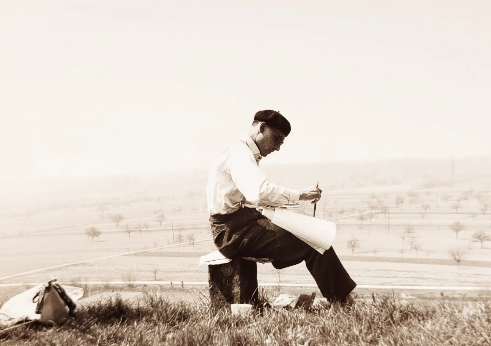Mann im Profil mit Pinsel und Zeichenpapier sitzt in einer weiten Landschaft.