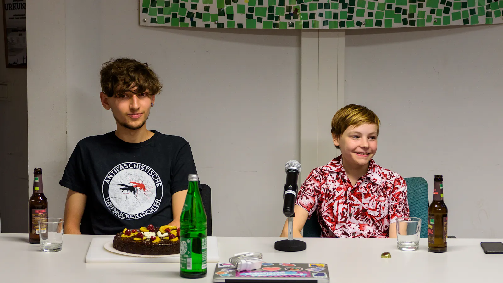 Jason und Cito sitzen am Tisch. Vor Jason steht eine vegane Torte mir einer 18. Auf dem Tisch stehen noch leere Malzbierflaschen, eine Wasserflasche, sowie Jasons geschlossener Laptop.