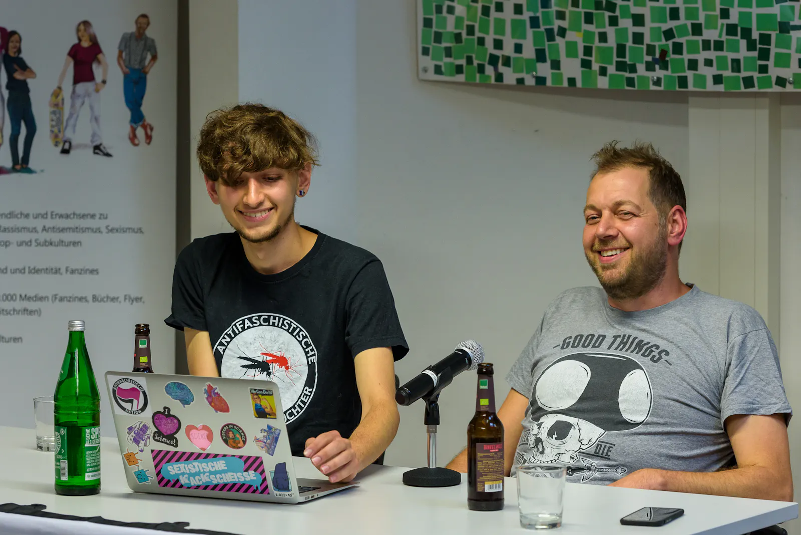 Jason und Mirco sitzen an einem Tisch, vor Ihnen ein Standmikrofon, Jasons Laptop, bestückt mit zahlreichen Aufklebern, Malzbier und Wasserflaschen.