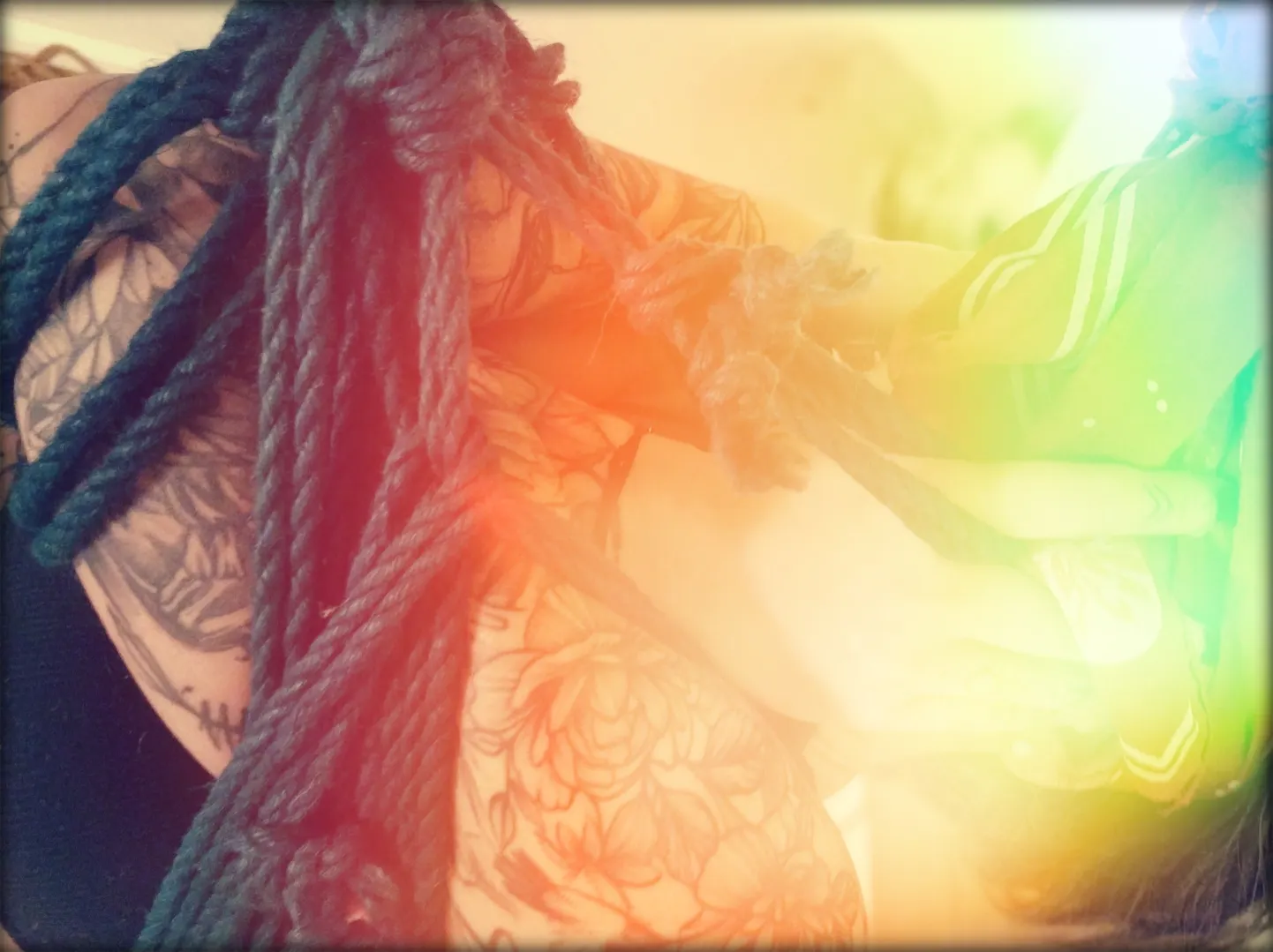 Von der Seite sehr nah zu sehen: tätowierte Arme, die den eigenen Oberkörper umschlingen - was wiederum in Gänze von Seilen umschlungen wird. Das bild hat eine Art Regenbogen-Farbverlauf als Lensflare Effekt.