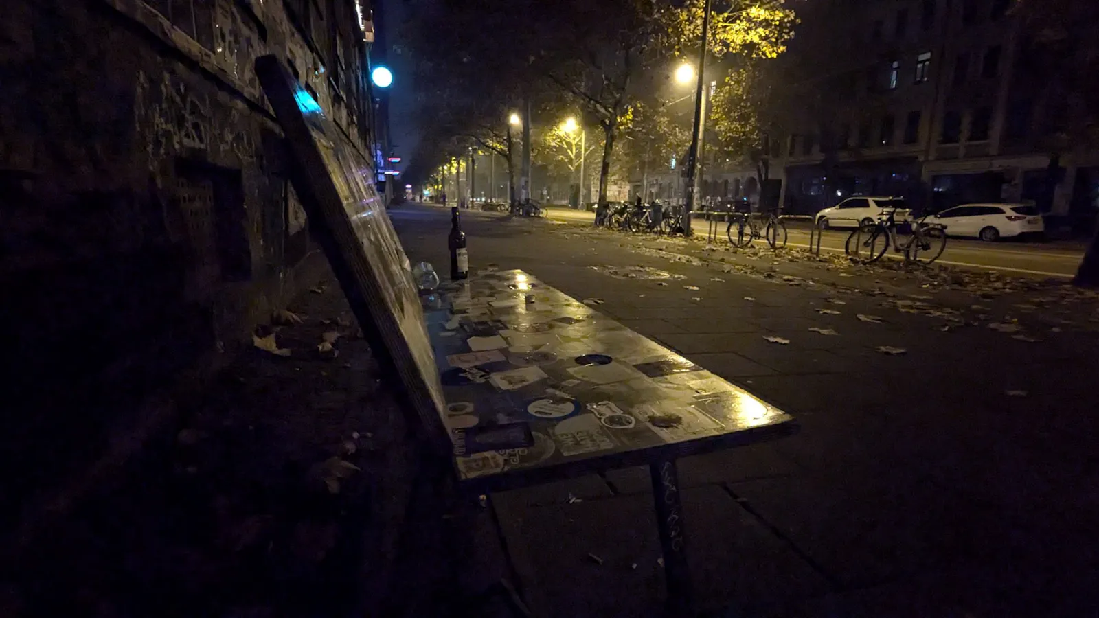 Eine nächtliche Straße, von Straßenlaternen beleuchtet. Im Vordergrund eine Bank, auf der eine leere Flasche steht.