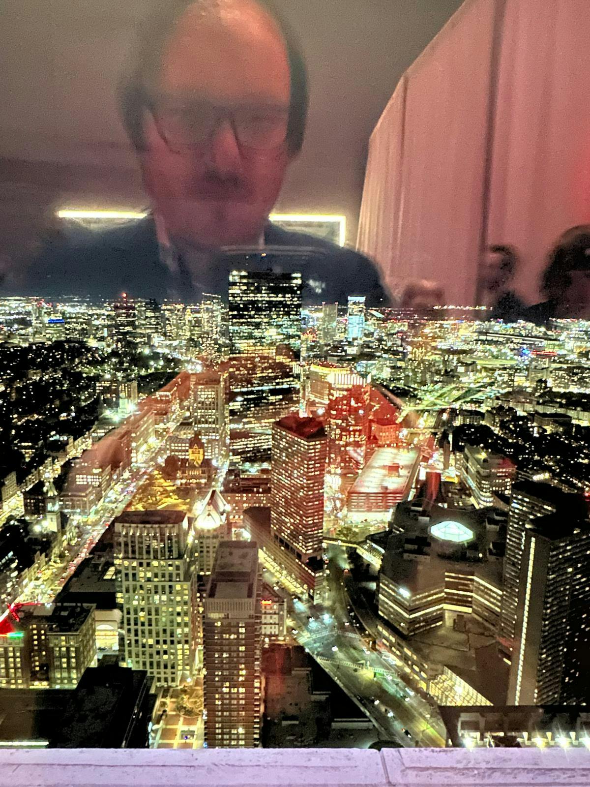 Es ist die erleuchtete Stadt Boston zu sehen, nachts, fotografiert aus einem Fenster des Prudential Towers. In der Spiegelung bin ich, Daniel Drepper, zu erkennen, wie ich das Foto mache.