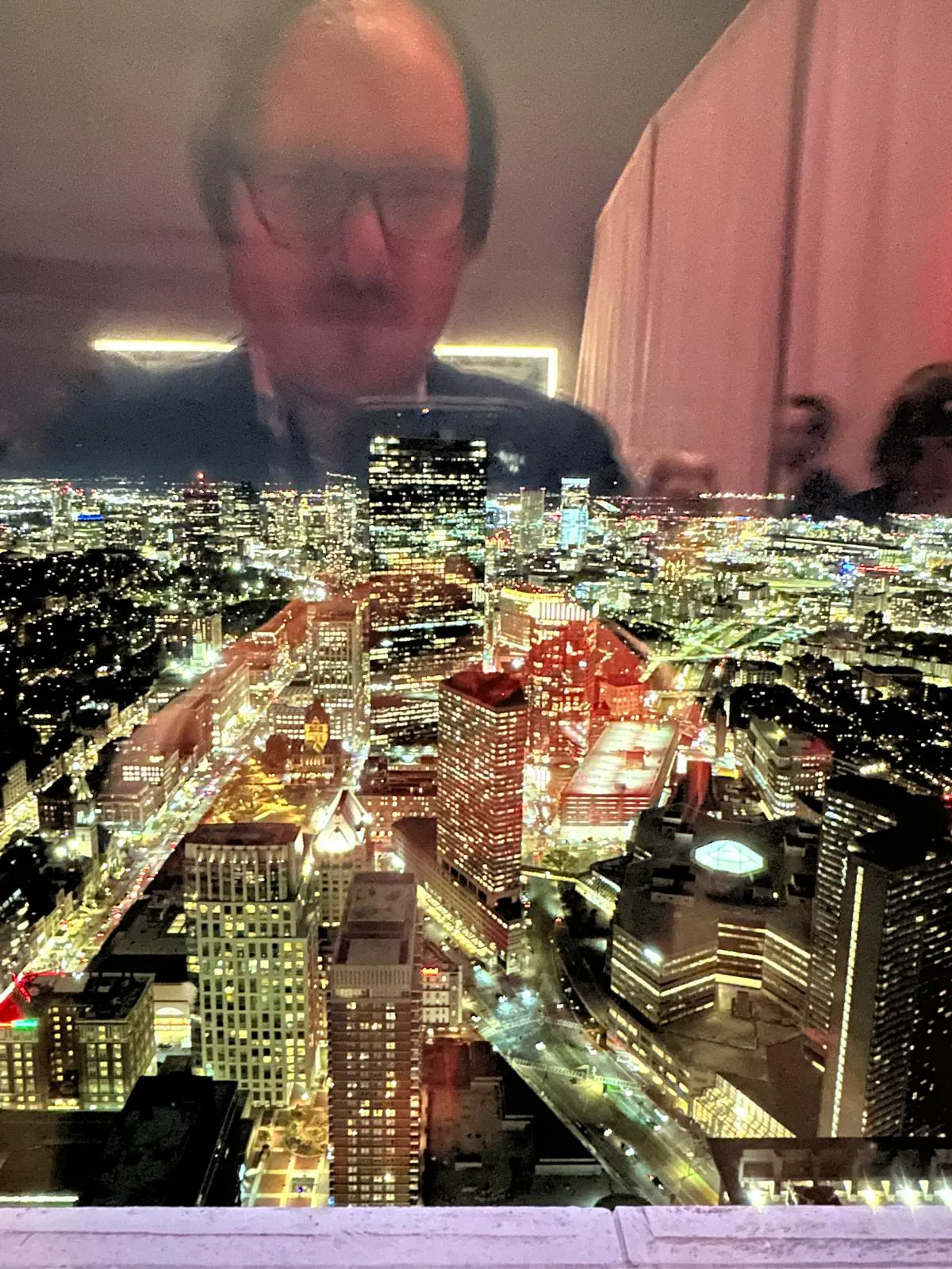 Es ist die erleuchtete Stadt Boston zu sehen, nachts, fotografiert aus einem Fenster des Prudential Towers. In der Spiegelung bin ich, Daniel Drepper, zu erkennen, wie ich das Foto mache.