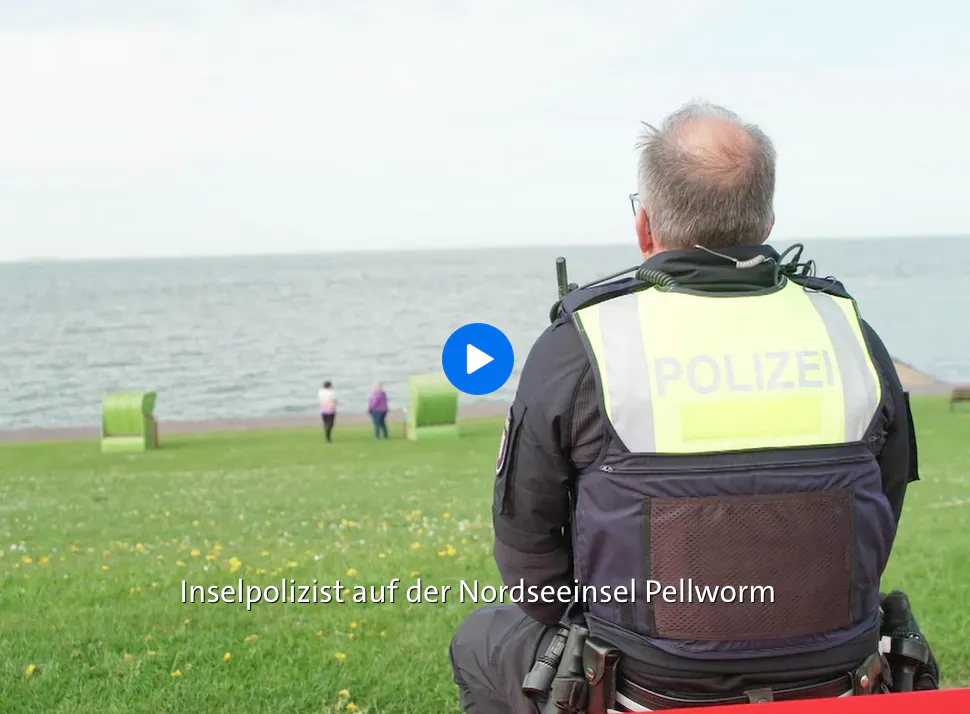 Markus in Uniform von hinten am Wasser. Man sieht Wiese, zwei Menschen klein weit hinten an der Wasserkante, zwei grüne Strandkörbe.