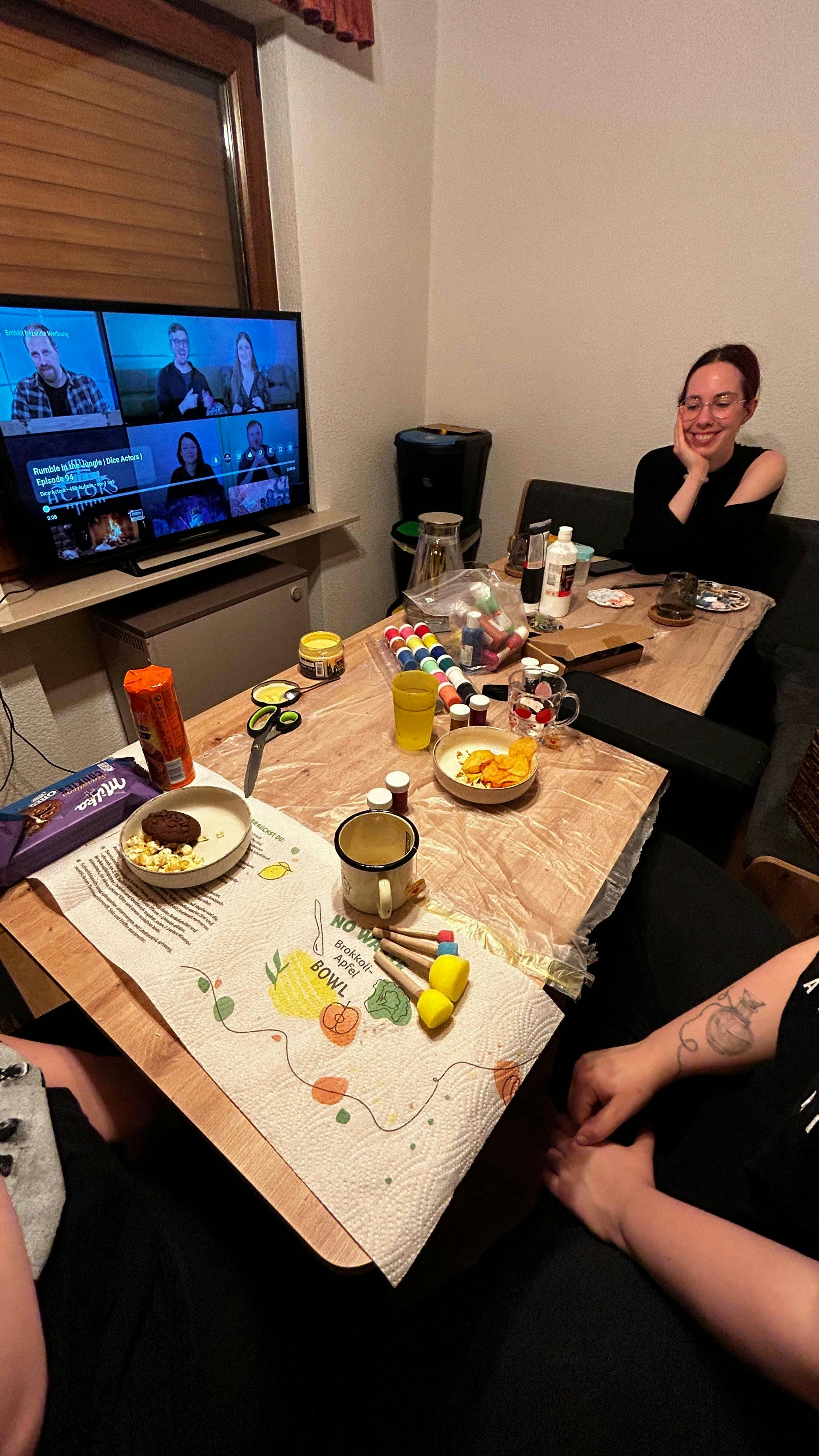 Auf einem Tisch stehen Farben und andere kreative Utensilien bereit. AM Fernseher im Hintergrund sieht man die Folge "Rumble in the Jungle".