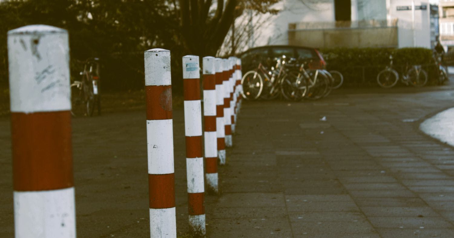 Blick auf rot-weiße Poller, im Hintergrund ein Haus und Fahrräder davor.