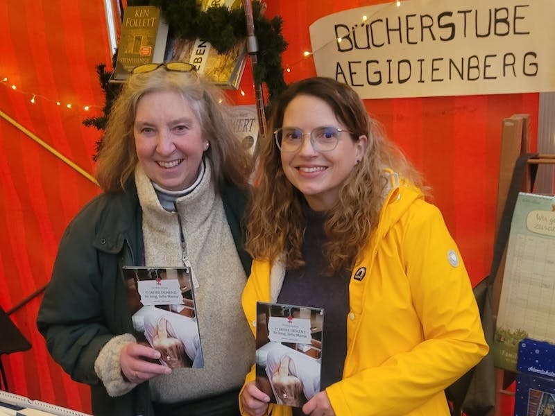 Verena Groß, Inhaberin der Bücherstube Aegidienberg, mit der Autorin Christine Piontek am Stand der Bücherstube auf dem Weihnachtsmarkt. Beide Frauen halten ein Exemplar des Buches "17 Jahre Demenz – So long, liebe Mama" von Christine Piontek hoch.