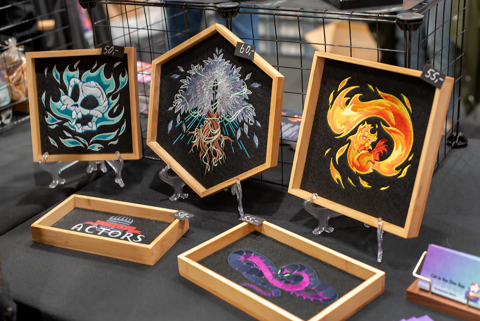 Eine Ăbersicht der Dice Trays: Gynther, Silberbaum, Fridolin, Dice-Actors-Logo und die VerhĂŒllte Schlange auf dem Verkaufstisch der AnimagiC 2025.