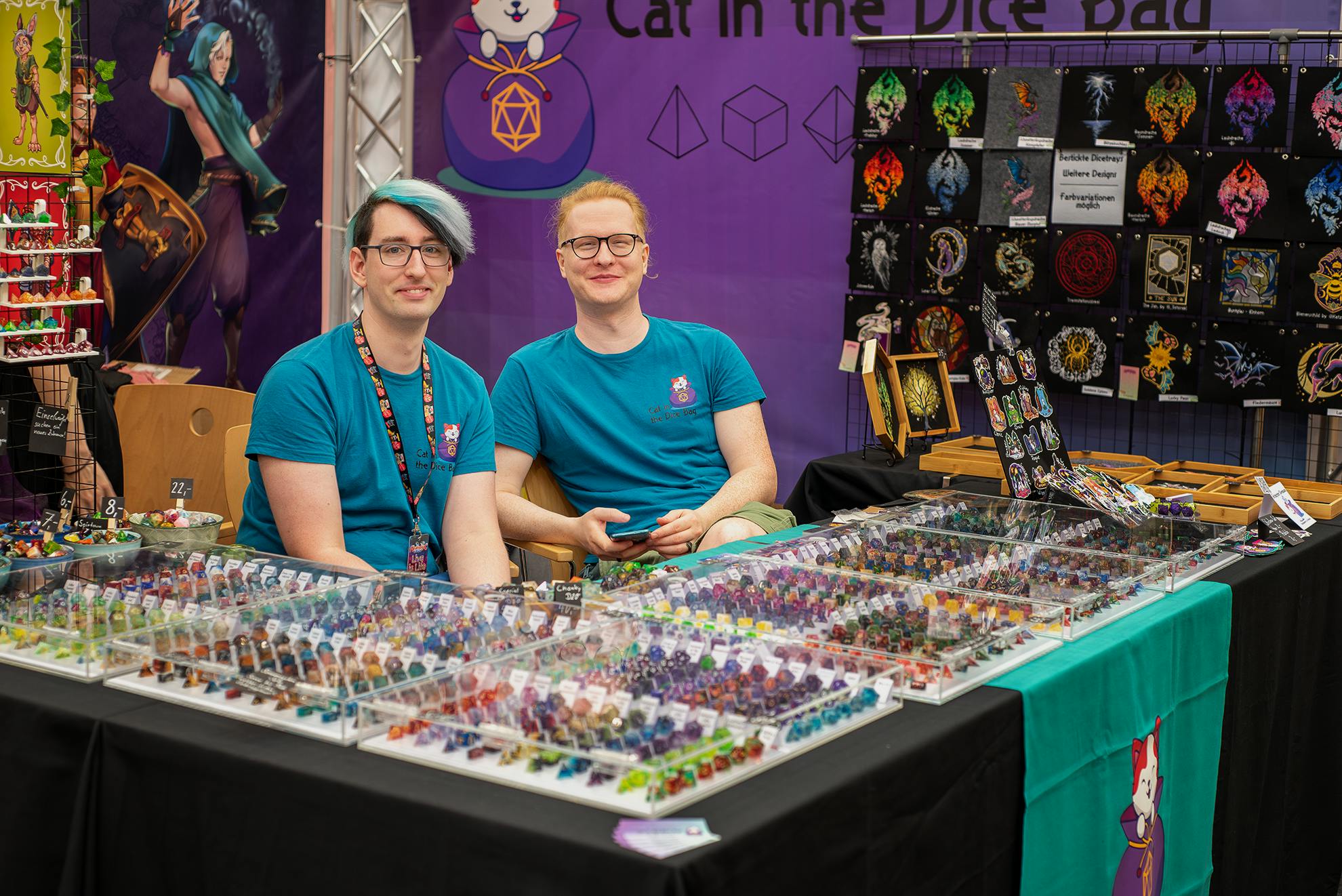 Philipp und Daniel von Cat in the Dice Bag an ihrem Stand auf einer Messe. Vor ihnen liegt hinter Glasabdeckungen ordentlich sortiert eine unzählbare Anzahl wunderschöner Würfel, die ich, Patze, alle sofort kaufen würde. Aber dann wäre ich vermutlich schnell pleite... es sind so, so, so viele Würfel...