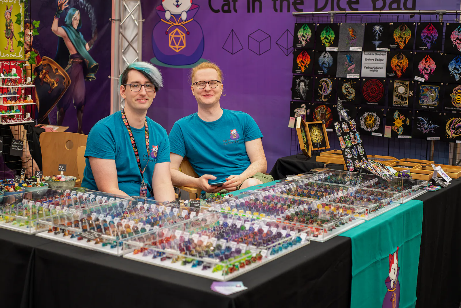 Philipp und Daniel von Cat in the Dice Bag an ihrem Stand auf einer Messe. Vor ihnen liegt hinter Glasabdeckungen ordentlich sortiert eine unzĂ€hlbare Anzahl wunderschöner WĂŒrfel, die ich, Patze, alle sofort kaufen wĂŒrde. Aber dann wĂ€re ich vermutlich schnell pleite... es sind so, so, so viele WĂŒrfel...