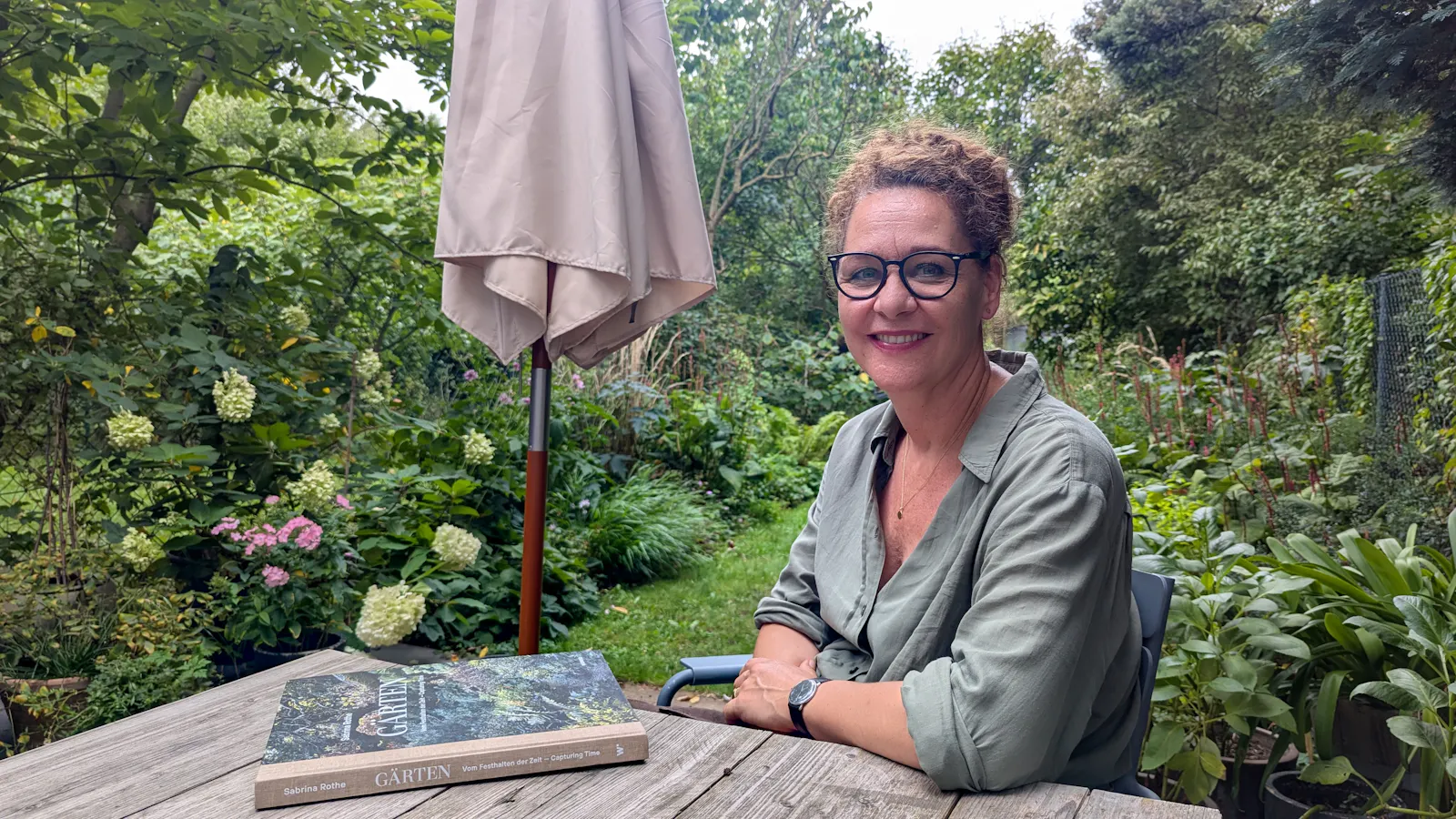 Sabrina Rothe sitzt an einem Holztisch im Garten, vor ihr ihr Buch „Gärten – Vom Festhalten der Zeit".