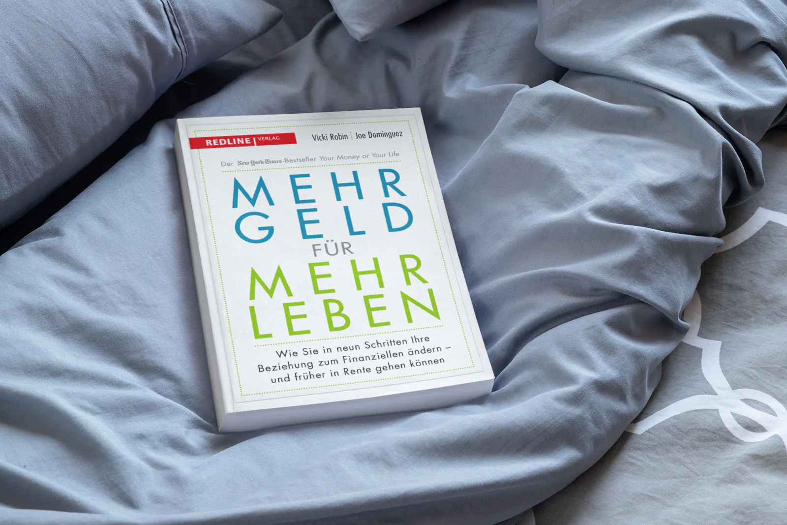 Ein Buch mit dem Titel "MEHR GELD FÜR MEHR LEBEN" liegt auf grauer Bettwäsche.