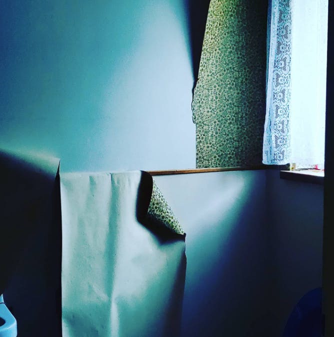 Detailaufnahme einer Zimmerecke mit heruntergerissenen, herunterhängenden Tapeten und einem Stück Spitzenvorhang vor dem Fenster, das man im Anschnitt sieht.
Das Foto ist in blau und grüntönen gehalten.