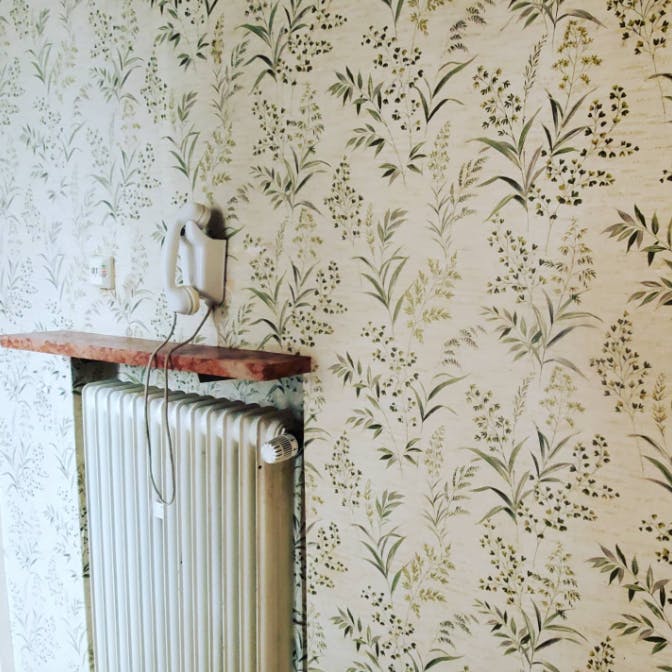 Detailaufnahme einer Wand mit Grastapete. Über einem Heizkörper in einer Nische ist eine Art Fensterbank montiert, darüber ein  weißes Festnetztelefon an die Wand montiert