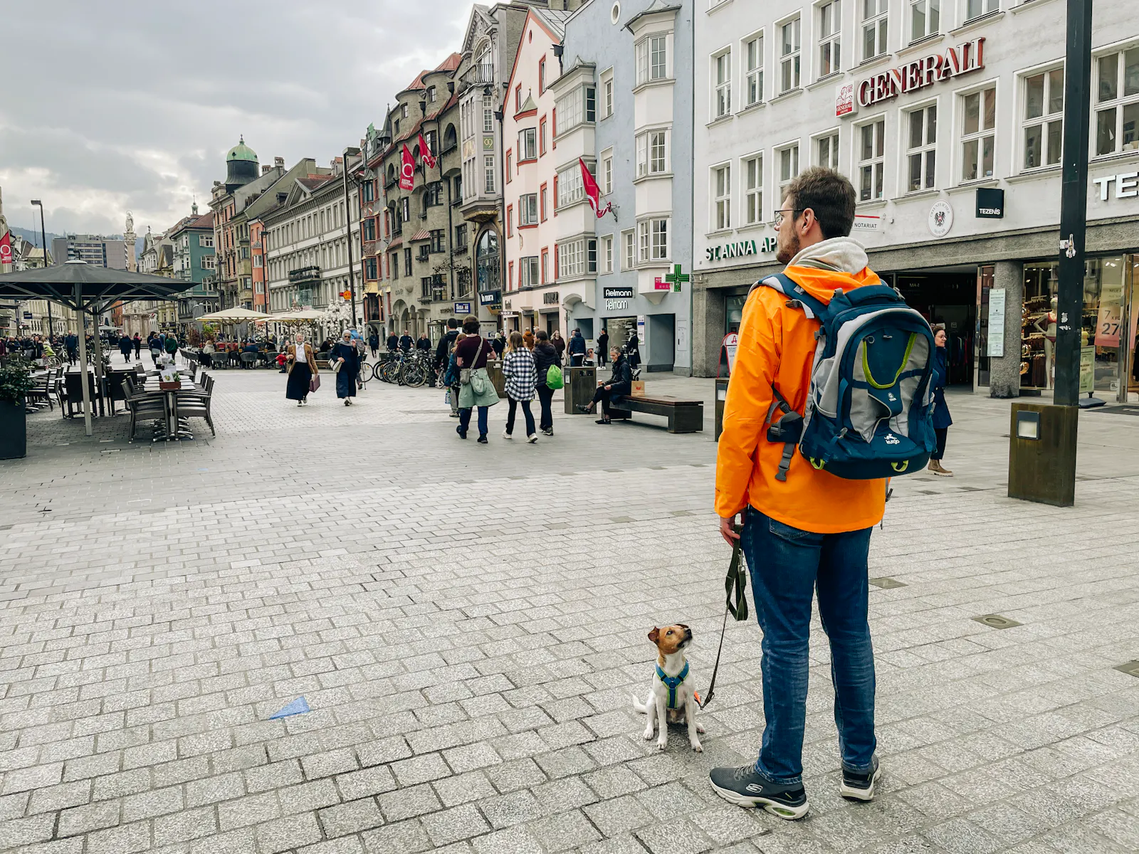 Pascal Turin und Redaktionshund Waldo stehen in Innsbruck auf der Maria-Theresien-Strasse. Waldo ist an der Leine und schaut zu Pascal hoch. Pascal trägt eine orange Regenjacke und einen Wanderrucksack.