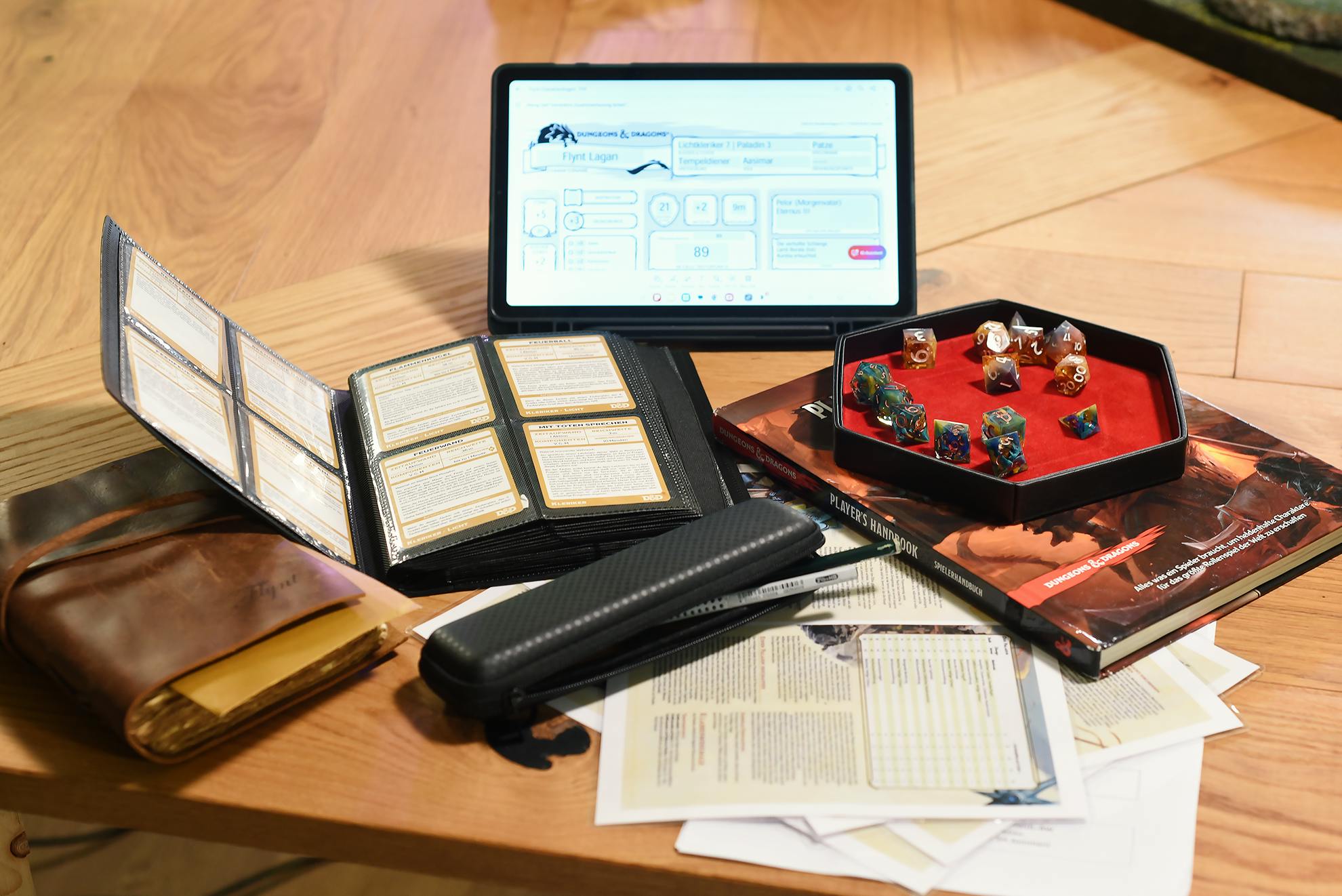 Ein Blick auf Patzes Spieltisch: Auf einem Holztisch liegt das komplette D&D-Equipment ausgebreitet. Links ein geschlossenes braunes Gebetsbuch aus dem ein goldener Umschlag herauslugt, daneben ein geöffneter Zauberkarten-Ordner mit mehreren eingesteckten Karten. In der Mitte verteilt liegen ausgedruckte Regelblätter und ein schwarzes Mäppchen. Rechts liegt das D&D-Spielerhandbuch, darauf ein schwarzes Dice Tray mit rotem Samteinleger und bunten Würfeln. Im Hintergrund steht ein Tablet mit dem digitalen Charakterbogen von Flynt Lagan.