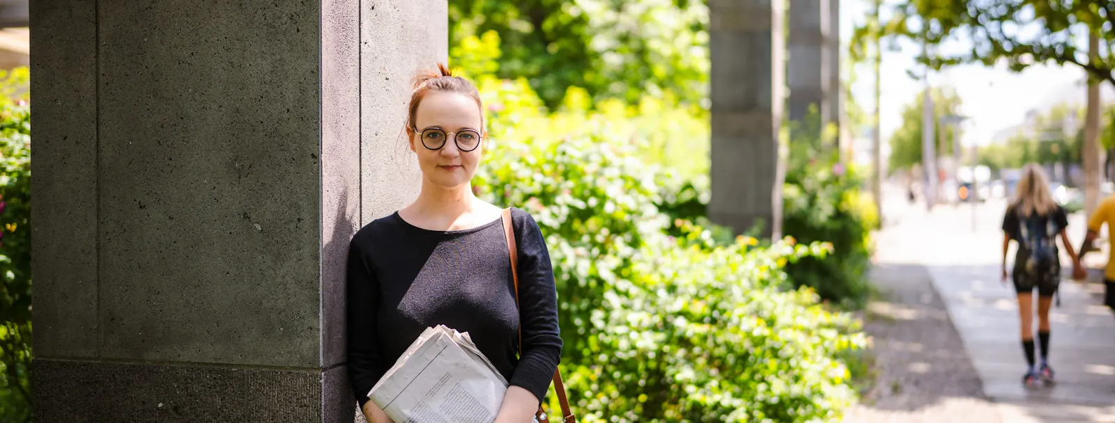 Insa van den Berg, Frau mit roten Haaren und Brille, lehnt mit einer Zeitung an einer Säule.