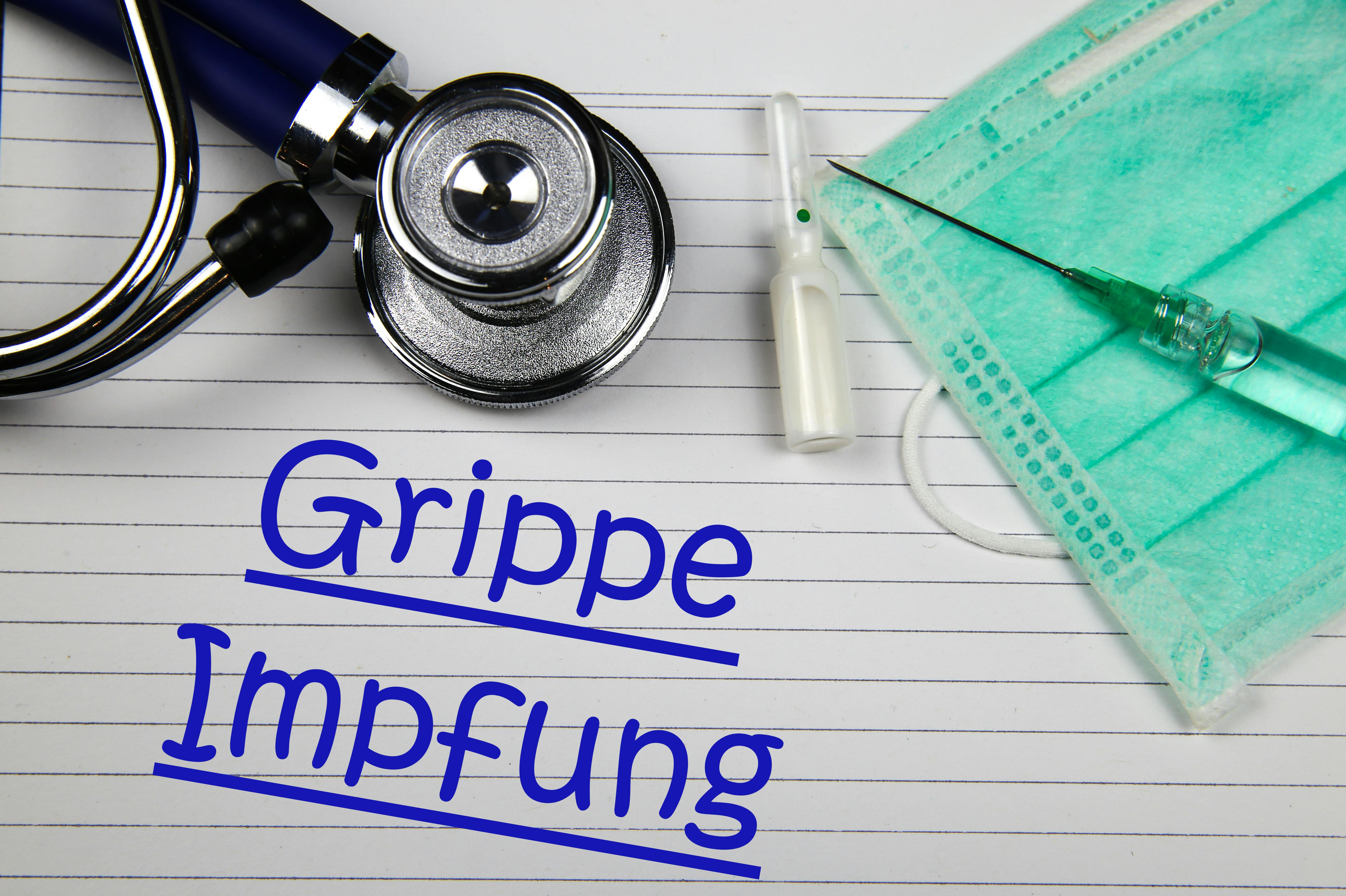 Das Bild zeigt ein Stethoskop, Einmalspritze mit Flüssigkeit und OP-Maske liegen auf einem linierten Blatt mit dem Schriftzug „Grippe Impfung“