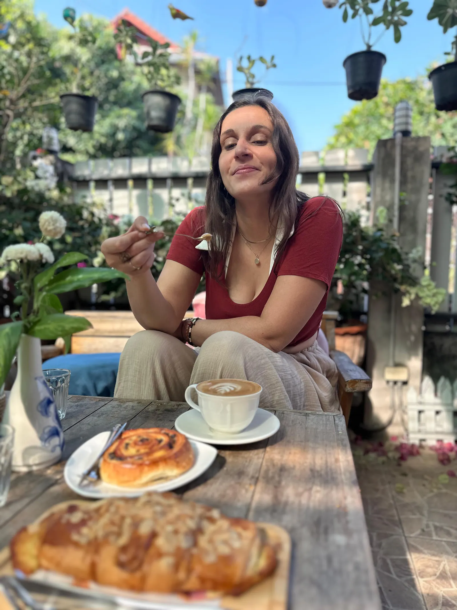 Josi sitzt lächelnd vor einem Tisch mit Kaffee und Kuchen in einem Café-Garten und hält einen Löffel in der Hand.