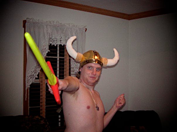 A photograph of me from 2007 dressed with a historically inaccurate horned Viking helmet in gold, bare chested with a silver necklace, and holding a yellow neon sword with a red hilt outward towards the camera in my right hand with my left hand behind me at an angle as though I am holding an invisible shield. This is Krowness the Skull Eater in his first ever appearance.