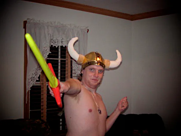 A photograph of me from 2007 dressed with a historically inaccurate horned Viking helmet in gold, bare chested with a silver necklace, and holding a yellow neon sword with a red hilt outward towards the camera in my right hand with my left hand behind me at an angle as though I am holding an invisible shield. This is Krowness the Skull Eater in his first ever appearance.