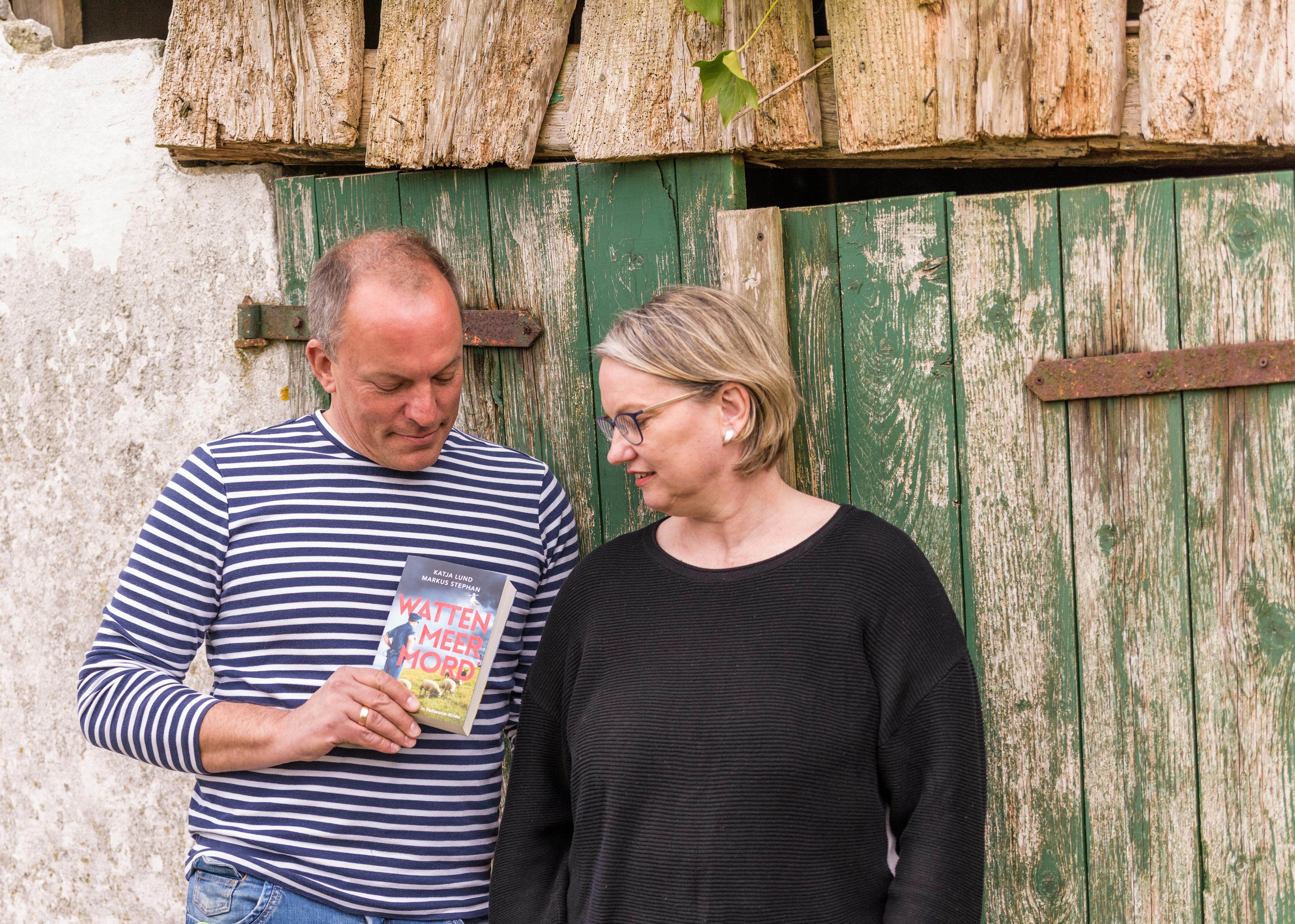 Markus Stephan und Katja Lund stehen vor einer grünen Holztür und betrachten ihr Buch "Wattenmeermord".