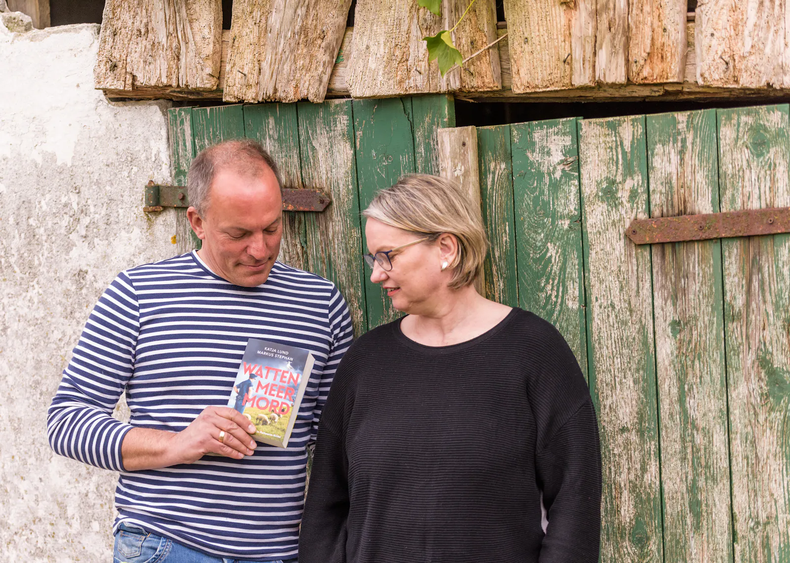 Markus Stephan und Katja Lund stehen vor einer grünen Holztür und betrachten ihr Buch "Wattenmeermord".