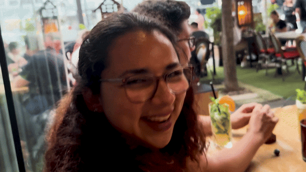 Gif-Datei: Aylin mit lockigem Haar und Brille lacht herzlich und klatscht sich begeistert in die Hände, weil sie sich auf ihr Essen freut. Sie sitzt mit den Dice Actors in einem Restaurant. Im Hintergrund sitzt Patze und sieht nach vorn zu Rieke, die nicht mehr mit im Bild ist. Vor Aylin steht ein Getränk mit einer Orangenscheibe.