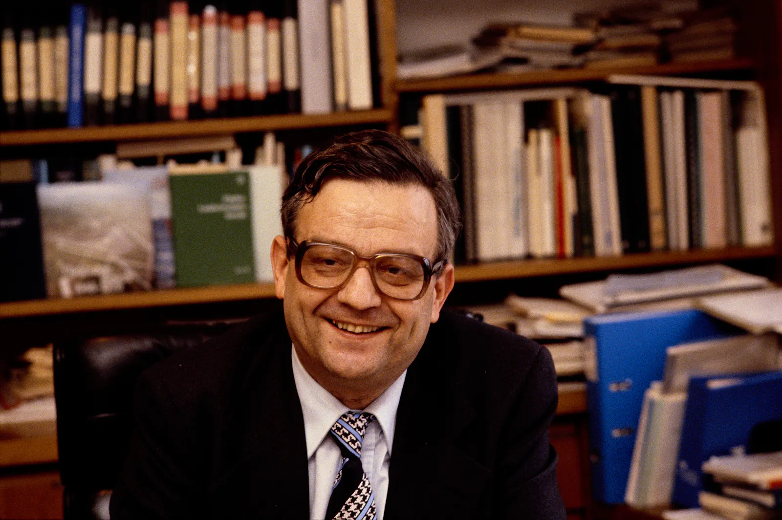 Hans Künzi sitzt 1971 mit Anzug und Krawatte in seinem Büro. Hinter ihm befindet sich ein Büchergestell voller Bücher. Hans Künzi trägt eine Brille und lächelt.