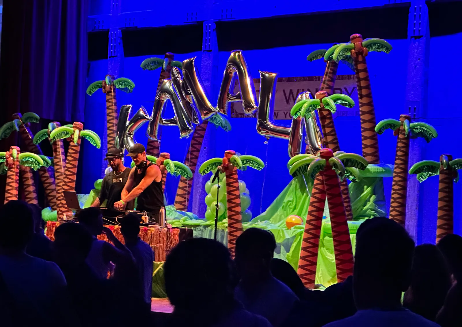 Zwei männliche DJs beim Event "Coconuts for Kamala", die vor aufgeblasenen Palmen und "Kamala"-Luftballons die Turntables bedienen.