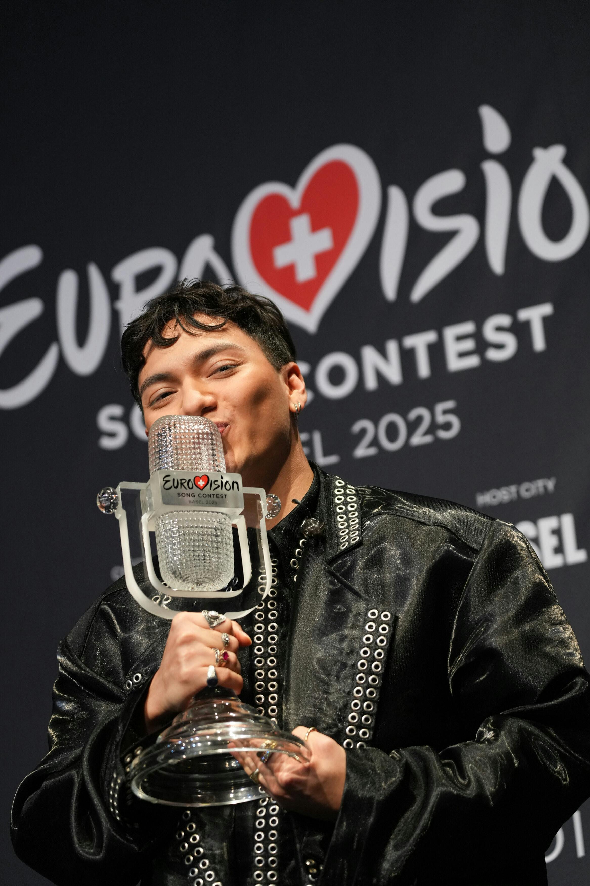 Der österreichische Sänger JJ hält die Eurovision Song Contest-Trophäe in der Hand und küsst sie. Im Hintergrund ist das Logo des Wettbewerbs mit der Aufschrift „Eurovision Song Contest 2025“ zu sehen.