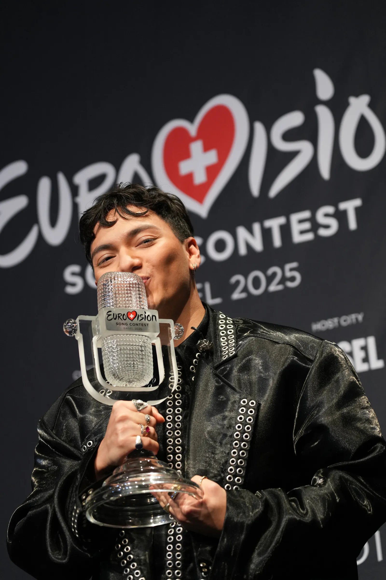 Der österreichische Sänger JJ hält die Eurovision Song Contest-Trophäe in der Hand und küsst sie. Im Hintergrund ist das Logo des Wettbewerbs mit der Aufschrift „Eurovision Song Contest 2025“ zu sehen.