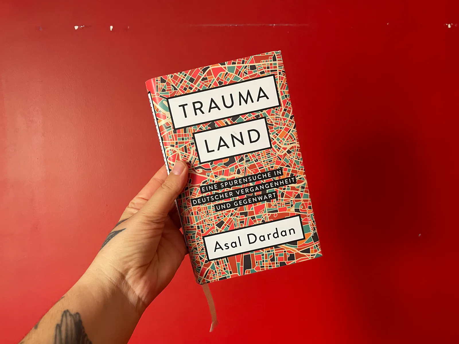 eine tätowierte hand hält das buch traumaland. eine spurensuche in deutscher vergangenheit und gegenwart von asal dardan vor einer roten wand an die kamera