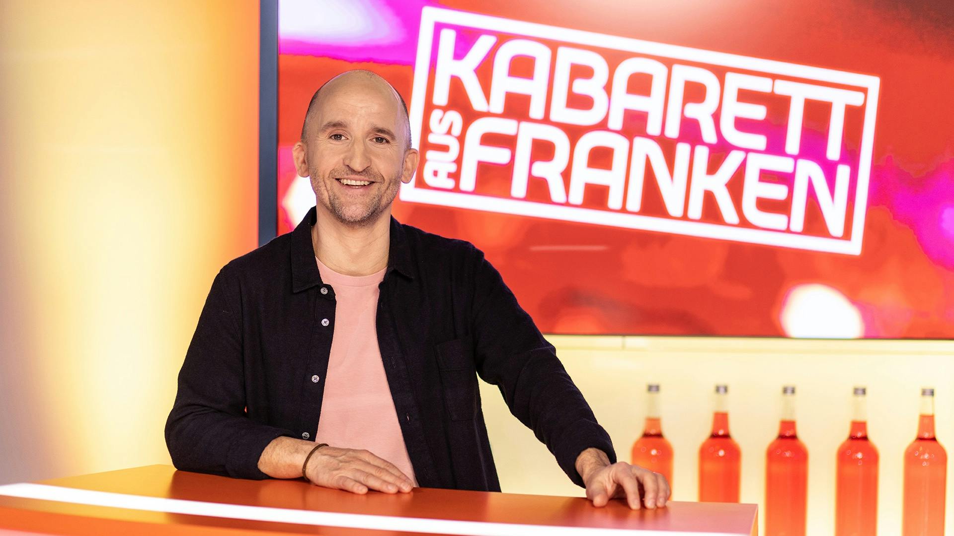 Ich in der Deko der TV Sendung Kabarett aus Franken. Hinter mir das Logo der Sendung auf einem Monitor, einige Flaschen mit farbiger Flüssigkeit. Ich stehe an einer Theke.