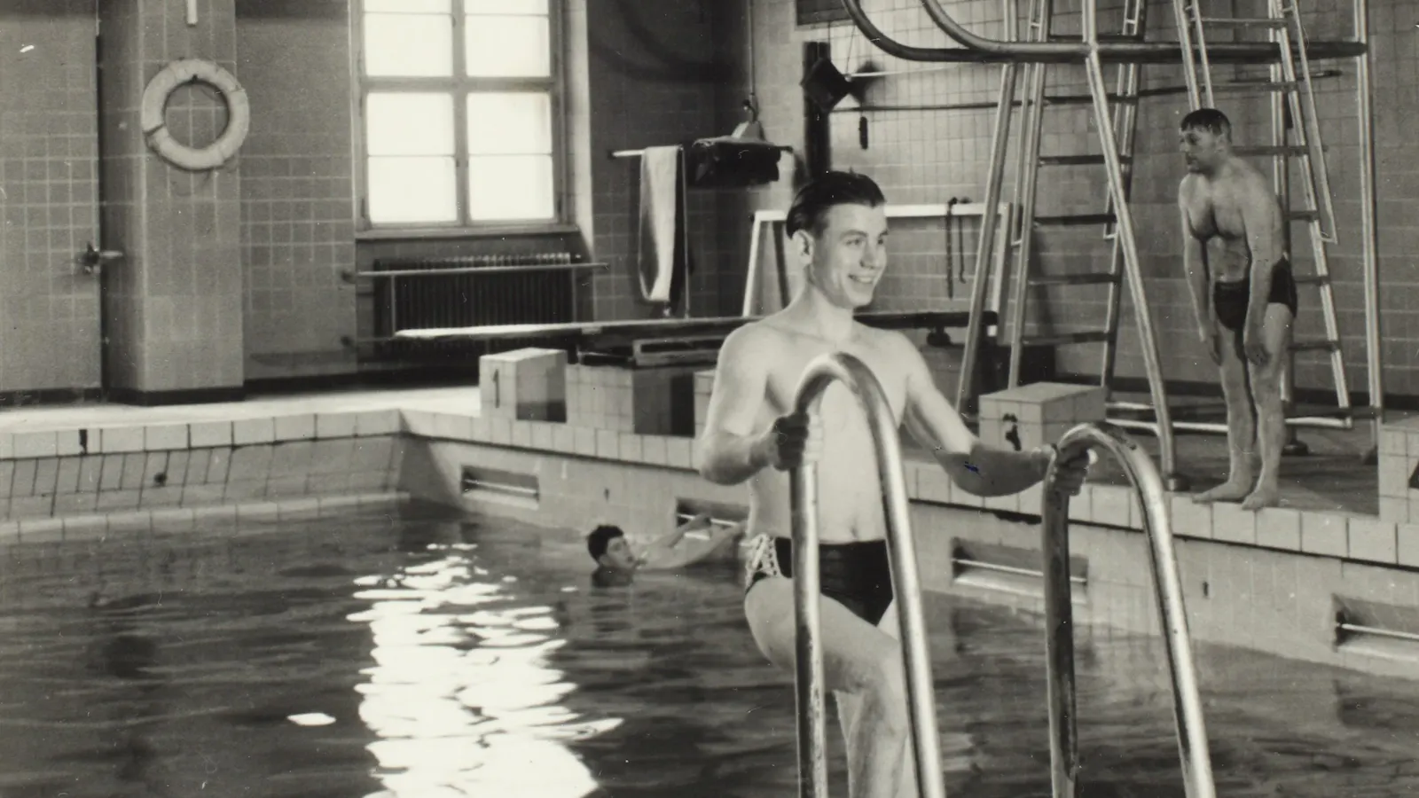 Schwarz-Weiß Foto von einem Schwimmbecken in einem Hallenbad. Ein junger Mann mit Badehose steigt gerade aus dem Wasser.