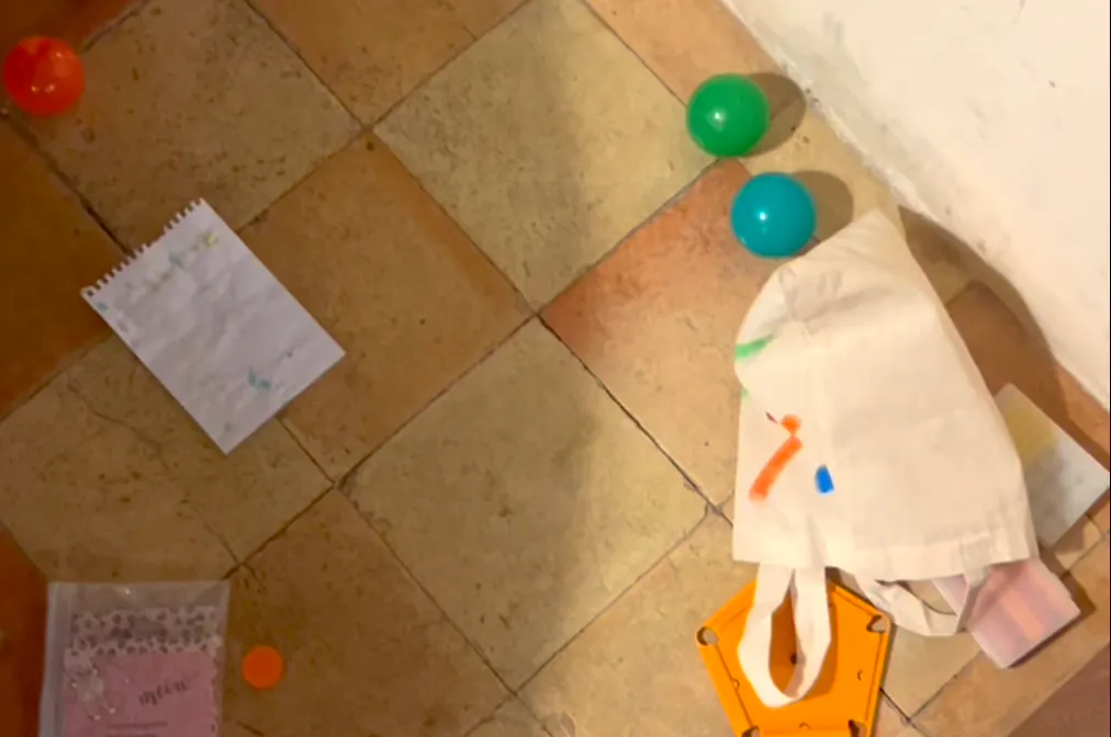 Das Bild zeigt einen Boden mit Fliesen in warmen Farben. Auf dem Boden befinden sich verschiedene Gegenstände: ein Blatt Papier, ein weißer Stoffbeutel mit bunten Farben, grüne, blaue und rote Bälle, ein orangefarbenes Plastikspielzeug.