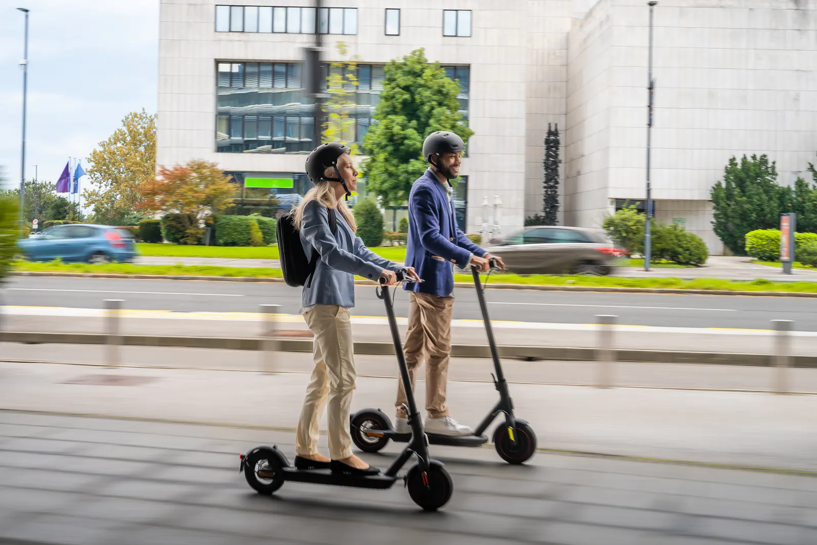 Das Bild zeigt eine Frau und einen Mann, die nebeneinander auf E-Scootern durch eine Stadt fahren. Beide tragen einen Helm.