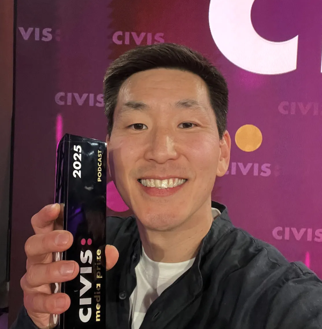 Frank macht ein Selfie, im Hintergrund ist das Logo des CIVIS Medienpreises. Er hält einen Preis in der Hand und lächelt.