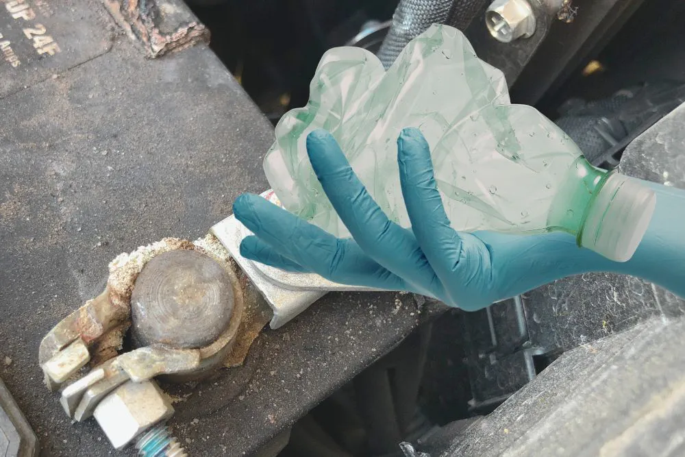 Une batterie au plomb à l'arrière-plan, avec une main gantée tenant un morceau de plastique au premier plan.
