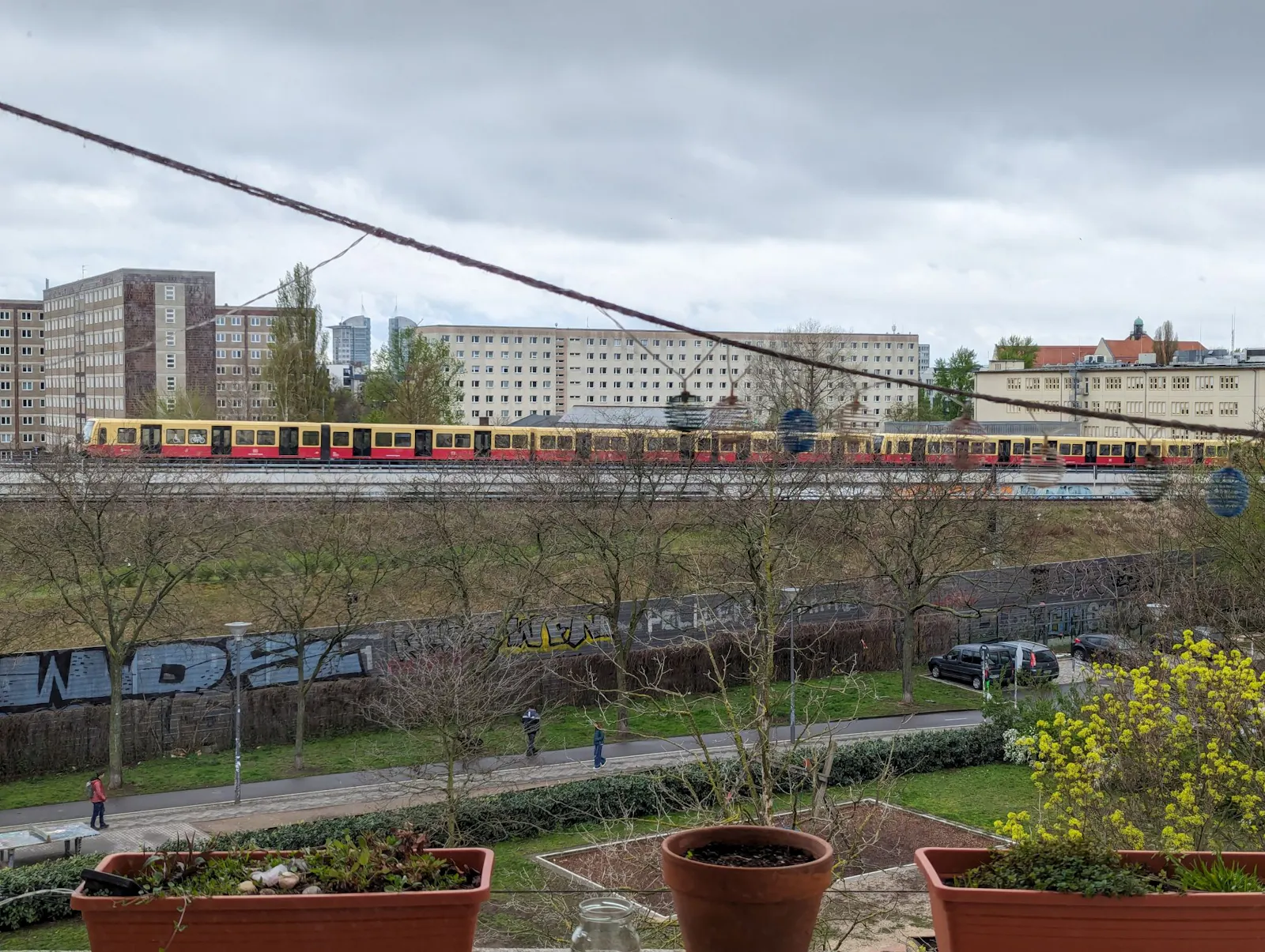 Im Hintergrund sieht man Plattenbauten, vor denen eine rotgelbe S-Bahn entlangfährt. Darüber graue Wolken. Im Vordergrund eine parkähnliche Anlage. Es könnte trostlos aussehen. Aber eigentlich sieht es ganz hübsch aus.