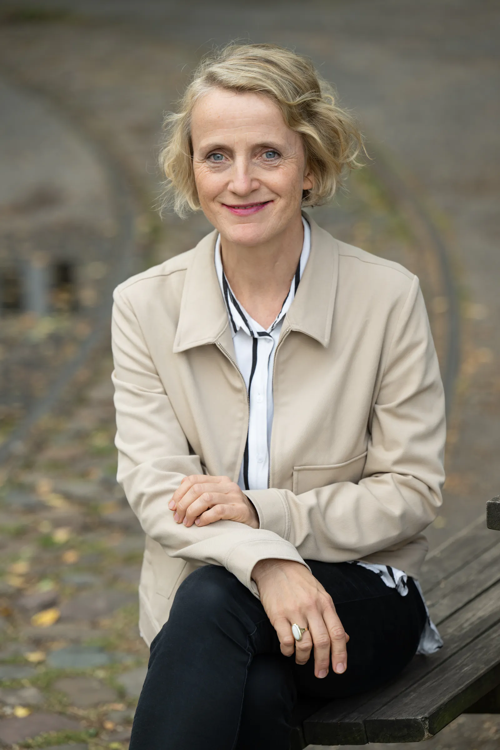 Eine Frau mit blonden, halblangen Haaren sitzt auf einer Holzbank. Sie trägt eine schwarze Hose und einen hellen Blazer.