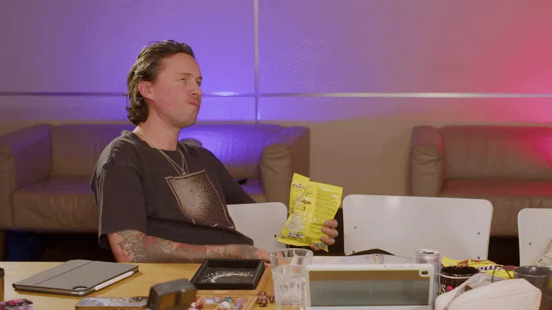 Patty sitzt am Spieltisch und hält eine gelbe Snacktüte in der Hand. Auf dem Tisch liegen Spielmaterialien wie Würfel, ein Tablet, Gläser und weiteres Zubehör. Im Hintergrund sind Sofas und farbiges Licht zu sehen. Patty versucht, sich einen der „Smash“-Chips in den Mund zu werfen, trifft aber nicht. Man sieht, dass er danach „Macht’s gut, ihr Idioten“ sagt.